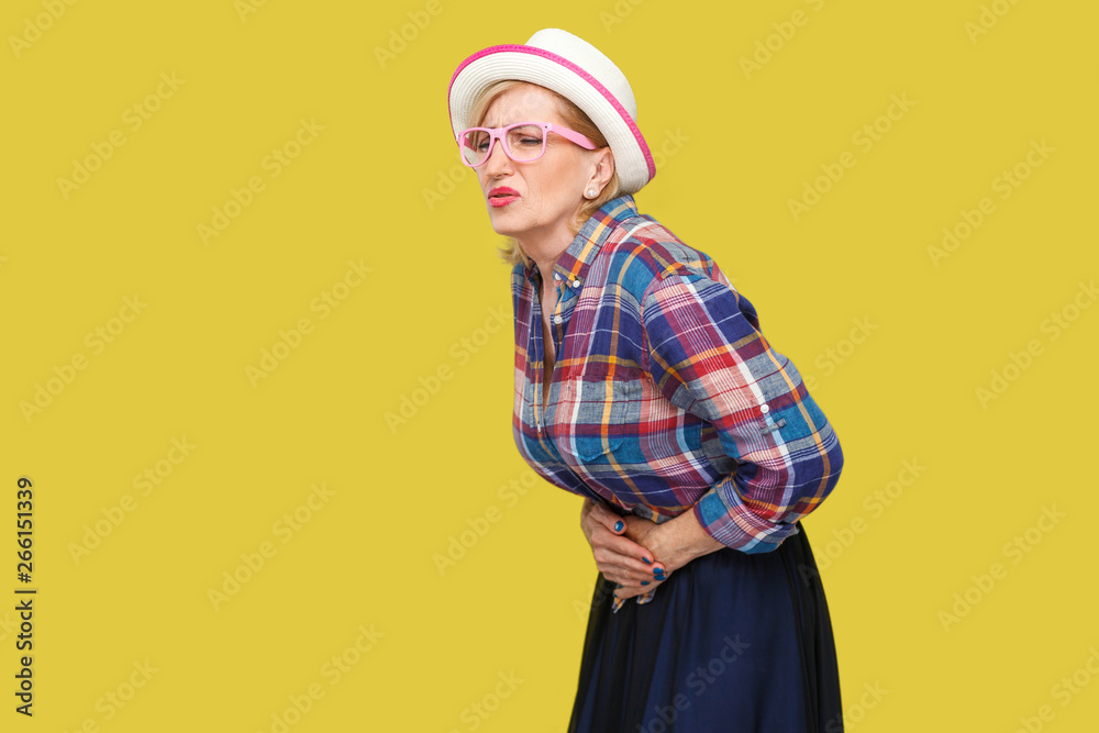 Stomach pain. Profile side view portrait of modern stylish mature woman in casual style with hat and eyeglasses standing and holding her painful belly. indoor studio shot isolated on yellow background