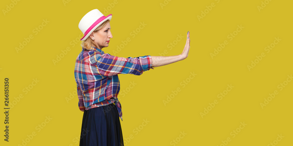 Stop it. Profile side view portrait of serious modern stylish mature woman in casual style with hat standing looking forward and showing stop sign. indoor studio shot isolated on yellow background.