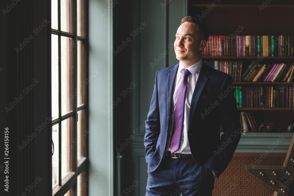 Fototapeta premium Portrait of a successful businessman standing in an expensive business suit near the window in his office and smiling