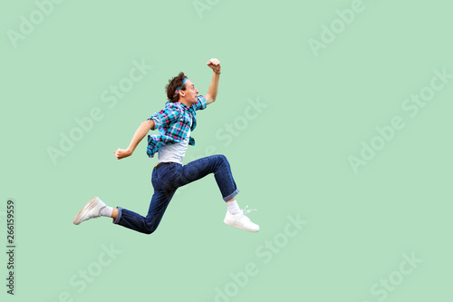 Jump to the success. Full length profile side view of active young man in casual blue checkered shirt and headband running very fast or jumping. indoor studio shot, isolated on green background. photo