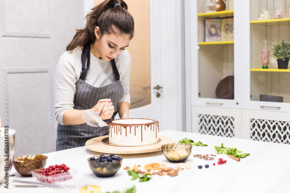 A confectioner squeezes liquid chocolate from a pastry bag onto a white cream biscuit cake on a wooden stand. The concept of homemade pastry, cooking cakes. A confectioner squeezes liquid chocolate from a pastry bag onto a white cream biscuit cake on a wooden stand. The concept of homemade pastry, cooking cakes.
