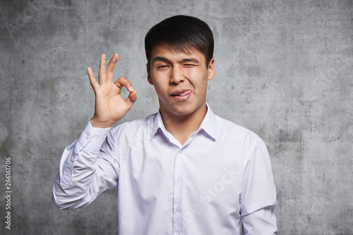 Young handsome Asian shows his fingers a gesture everything is OK. Joyful happy kazakh in business style clothes on gray background one