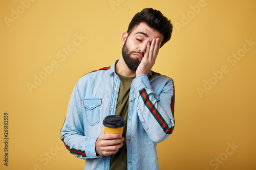 Tired exhausted student with sleepy expression, covers face with palm, holds disposable paper cup of coffee. man doesn't want to go to work. isolated yellow background