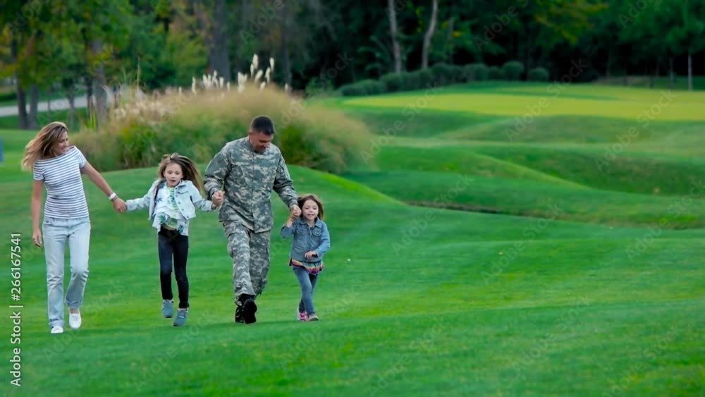 American family with father US soldier playing in the park lawn. Mother and father jumping with daughters on the meadow during day. American family with father US soldier playing in the park lawn. Mother and father jumping with daughters on the meadow during day.