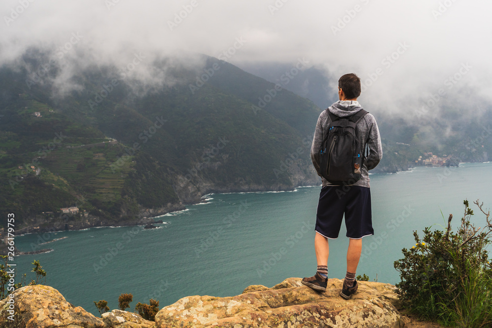 Male tourist wearing a hoodie, shorts and carrying a backpack admiring the beautiful view from the Punta Mesco where the Monterosso al Mare coastline meets the Ligurian Sea in Cinque Terre, Italy.
