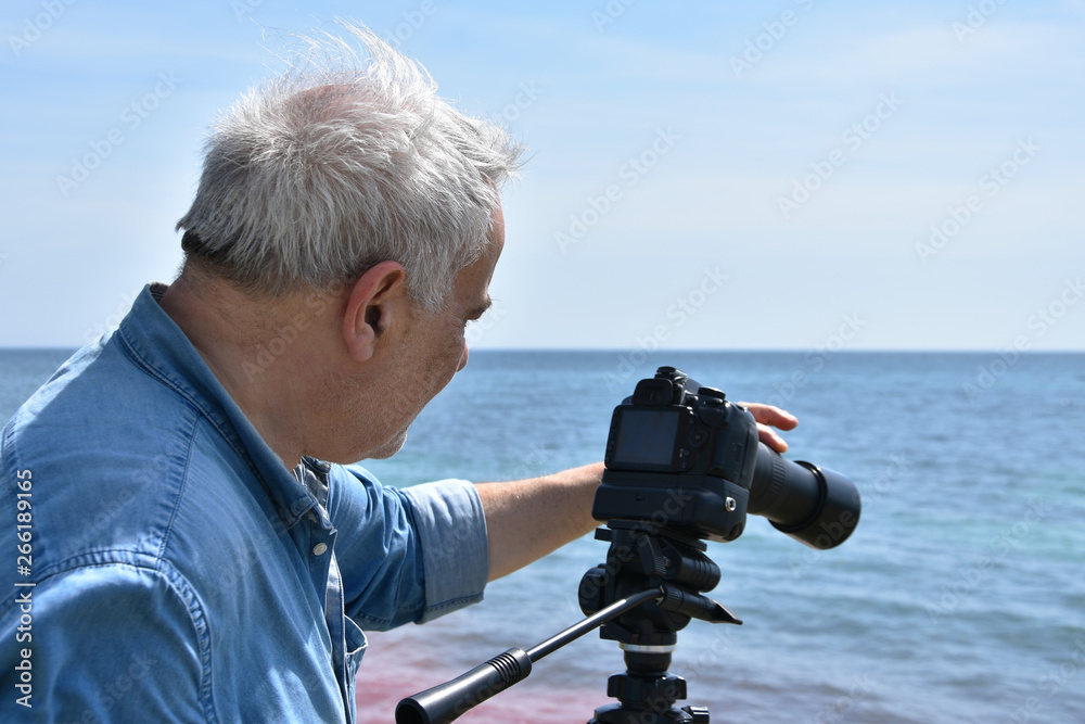 Obraz premium Portrait of an elderly man photographer by the sea. Senior adult shooting video with DSLR camera with telephoto lens on tripod.