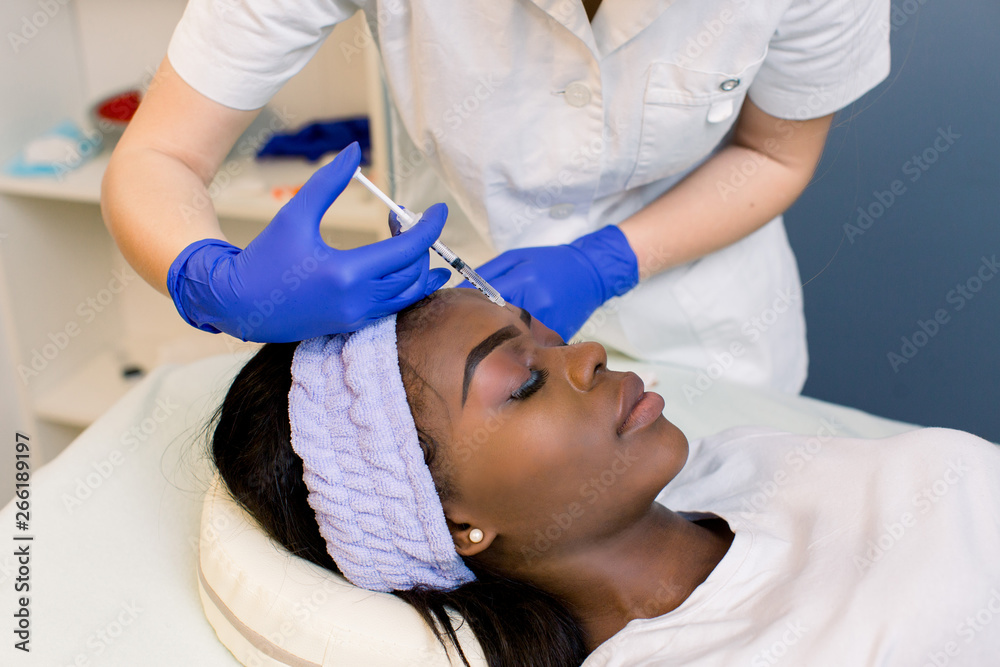 Close up of beautician expert's hands injecting botox in female forehead og young African girl. Close up of beautician expert's hands injecting botox in female forehead og young African girl.