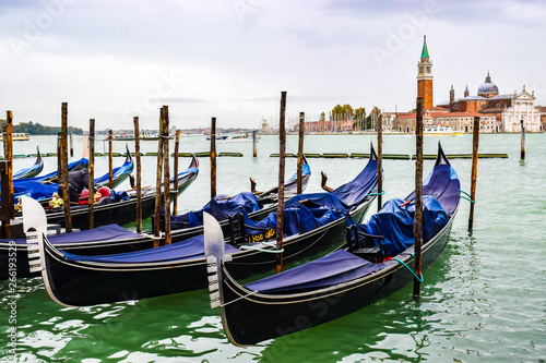 Covered gondolas docked on water between wooden mooring poles. Bell tower in background is of 16th century Palladian architecture Church of San Giorgio Maggiore on San Giorgio Maggiore island, Venice.