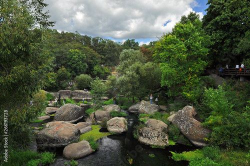 Huge boulders lie in the river among high green trees against the blue sky
