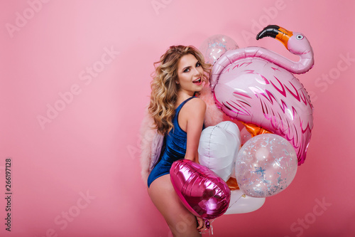 Cheerful curly girl in dark-blue velvet combination poses with a toy inflatable flamingo on pink background. Portrait of gorgeous blonde young woman holding colorful party balloons and having fun