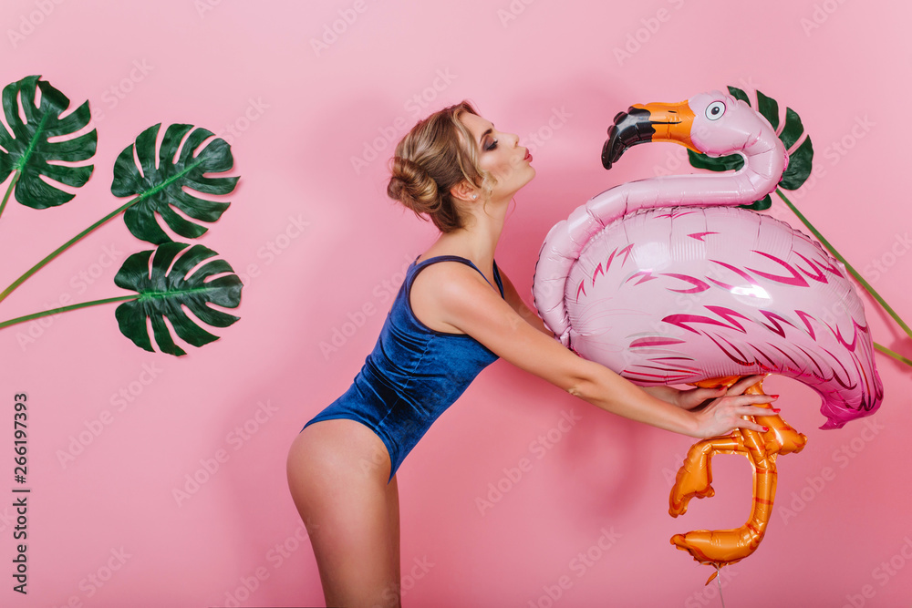 Slim amazing girl in vintage bodysuit kissing big toy bird, standing in  front of pink wall. Portrait of cute shapely young woman holding inflatable  flamingo, posing with plants on background Stock Photo