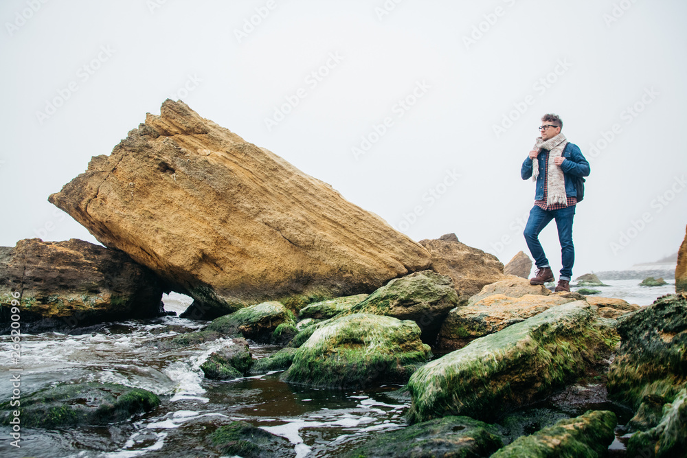 Man traveler with a backpack standing on a rock against a beautiful sea with waves, a stylish hipster boy posing near a calm ocean during a wonderful journey around the world