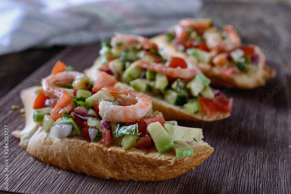 Appetizing bruschetta with a garnish of finely chopped mix of various fresh vegetables, avocados, decorated with shrimps on top, are on a wooden board