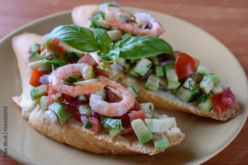 Appetizing bruschetta with a garnish of finely chopped mix of various fresh vegetables, avocados, decorated with shrimps on top, are on a plate