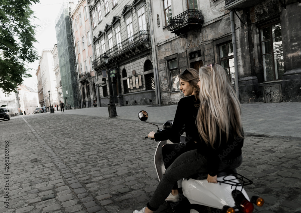 Fototapeta premium Young and beautiful girls in sunglasses riding a scooter. Portrait of pretty ladies riding a motorbike on old downtown street. Summer vacation. Girls in a black t-shirts on a vintage scooter