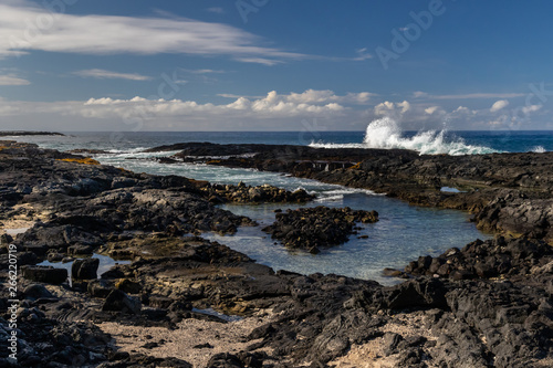 Tidepool at volcanic beach near Kona, on Hawaii's Big Island. Black rocks in foreground, with waves from incoming tide. Blue ocean, sky and clouds in the distance. 