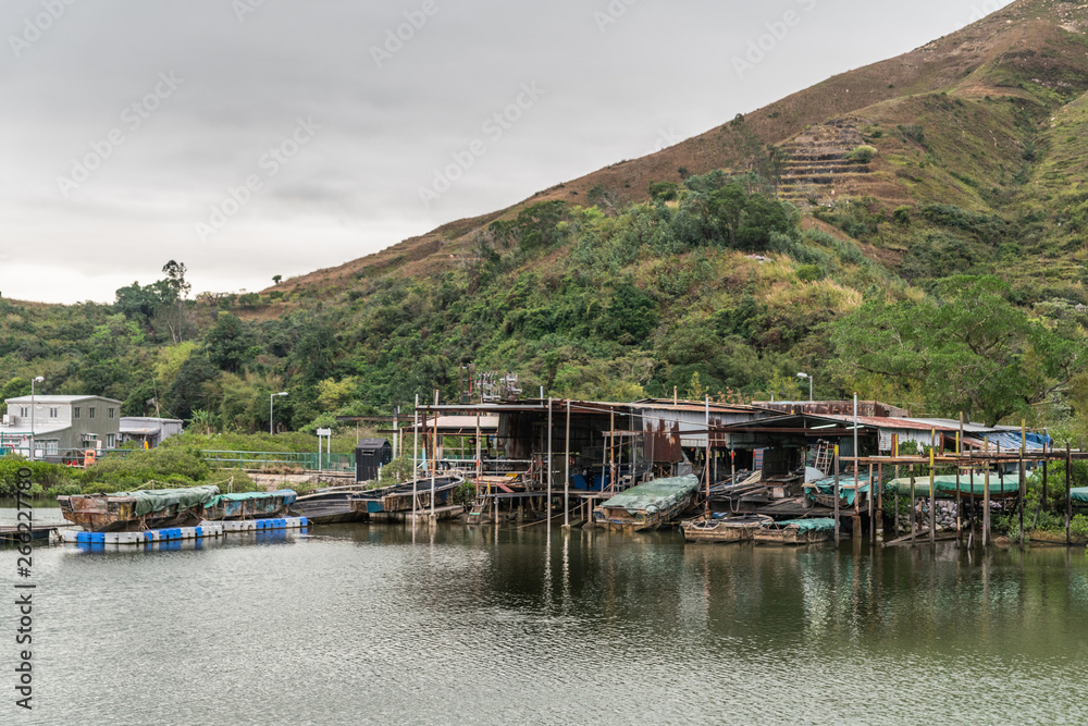 Hong Kong, China - March 7, 2019: Fishermen houses on stilts on Tai O River with large green hill in back under gray sky. Sloops, tarps and simple dwellings. Hong Kong, China - March 7, 2019: Fishermen houses on stilts on Tai O River with large green hill in back under gray sky. Sloops, tarps and simple dwellings.