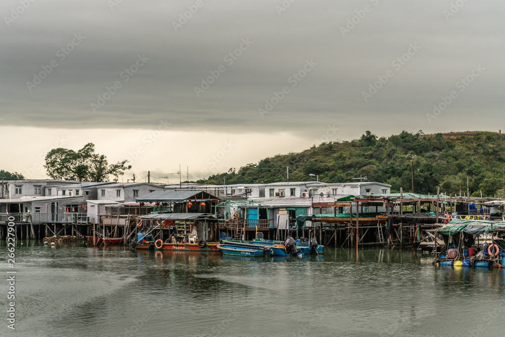 Hong Kong, China - March 7, 2019: White Fishermen houses on stilts on Tai O River with green hill in back under gray sky with yellow horizon. Color added by sloops, buoys and tarps.