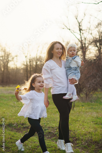 Happy young family on a walk in the spring meadow. Mom holds toddler in her arms. Girls in casual wear. Family look white clothes. Сaught at the moment scamper