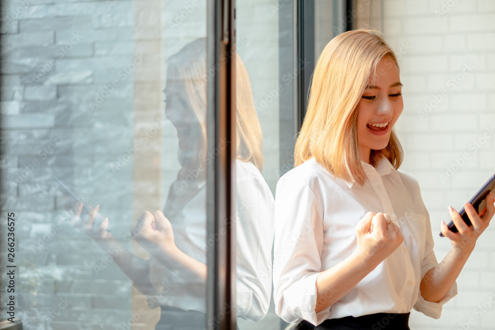 Fototapeta premium Young businesswoman standing near the office window smiling arms up celebrating with mobile phone success work at the office, business concept, success concept