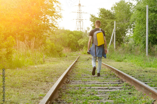 young man in jeans with backpack on his back goes forward along abandoned railway in summer sunset. concept of walking travel