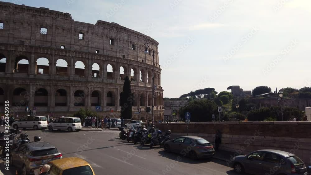 The colosseum from a different side than usual on a sunny spring day. There are few cars parked on the sides of the street. Cars are passing by. People are walking on the sidewalk. slow-motion.