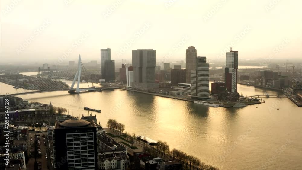 The eastern part of Rotterdam at sunrise. The iconic architecture is painetd in golden light. The river refelcts the sun and boats are cruising through the city of Holland.