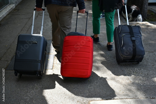Three suitcases, travel bags, on wheels. Two black and one red in the middle on the sidewalk of the city street. Visible hands of people pulling suitcases, legs and shadows. Travel, tourism, vacation. © Marijana