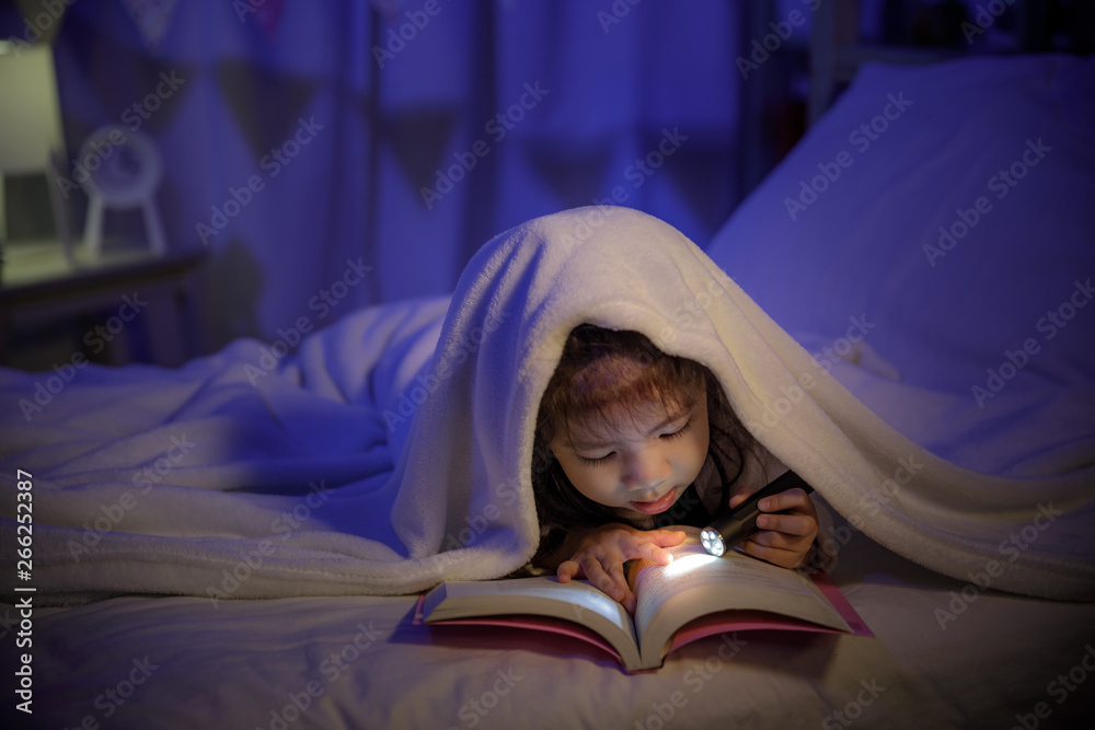 Child asian girl reading a book with flashlight under the blanket on bed in a dark bedroom at night before sleeping, Comfortable children at home concept Child asian girl reading a book with flashlight under the blanket on bed in a dark bedroom at night before sleeping, Comfortable children at home concept