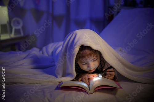 Child asian girl reading a book with flashlight under the blanket on bed in a dark bedroom at night before sleeping, Comfortable children at home concept