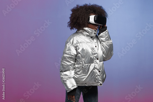 Trendy curly brown-haired girl dressed in a silver-colored jacket uses the virtual reality glasses in the studio on neon background