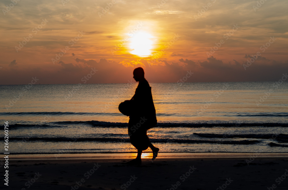 Sunrise with reflection on the sea and beach that have blurred silhouette photo of buddhist monk walking alms offering food in the morning on beach of Thailand.