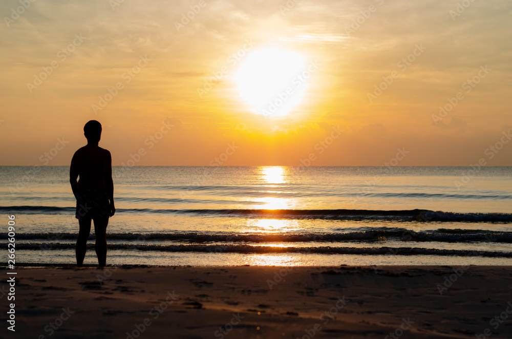 The silhouette photo of a man standing alone on the beach enjoy sunrise moment with the reflection on the sea in summer season.