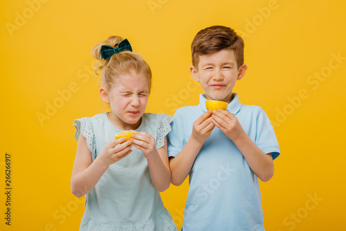 two young children, little girl and little boy taste sour lemon emotional, with sore, facial emotions negative, in blue T-shirt, isolated yellow background, copy space