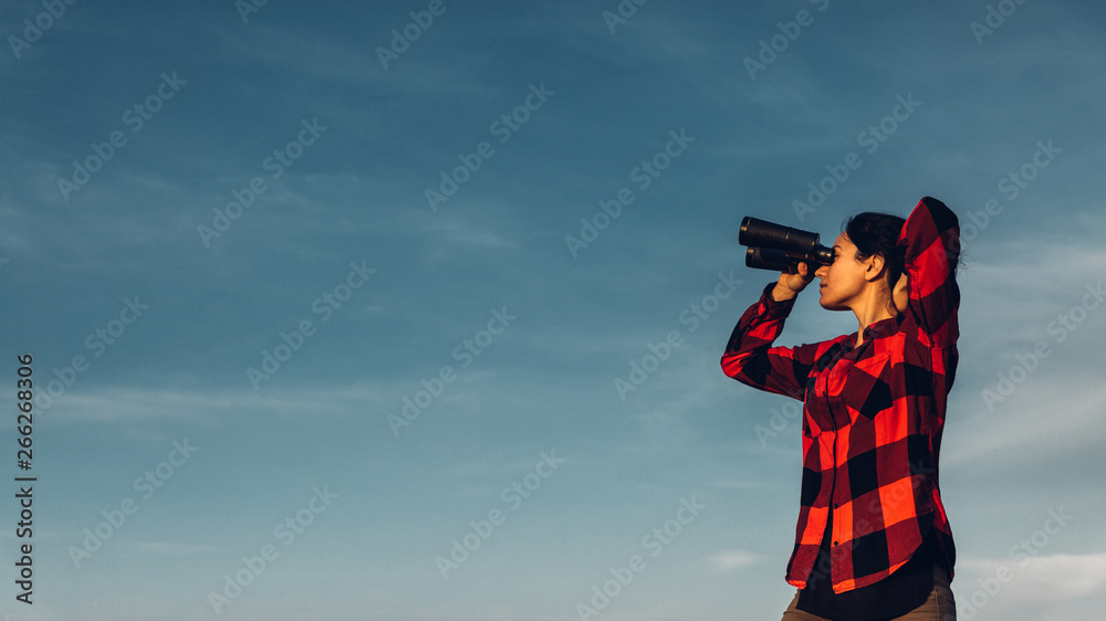 Obraz premium Beautiful young girl traveler looks through binoculars against a blue sky with copy-space. Concept of search, motivation, success, adventure