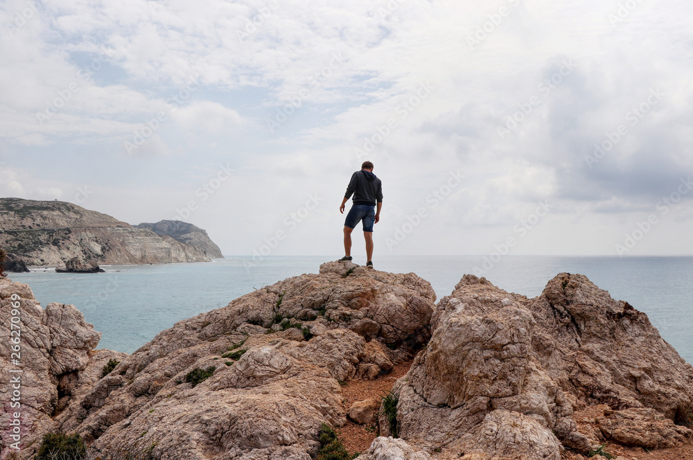 Young man stands on top of rock hill in the middle of mediterranean sea. View on ocean and cliffs in the distance. Famous and beautiful place in Cyprus nearby Kouklia. Petra tou romiou. Aphrodite rock