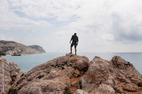 Young man stands on top of rock hill in the middle of mediterranean sea. View on ocean and cliffs in the distance. Famous and beautiful place in Cyprus nearby Kouklia. Petra tou romiou. Aphrodite rock