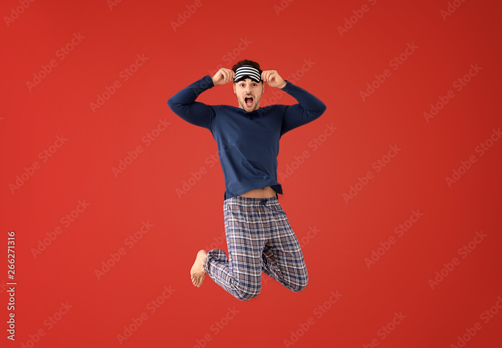 Jumping young man in pajamas with sleep mask on color background - obrazy, fototapety, plakaty 