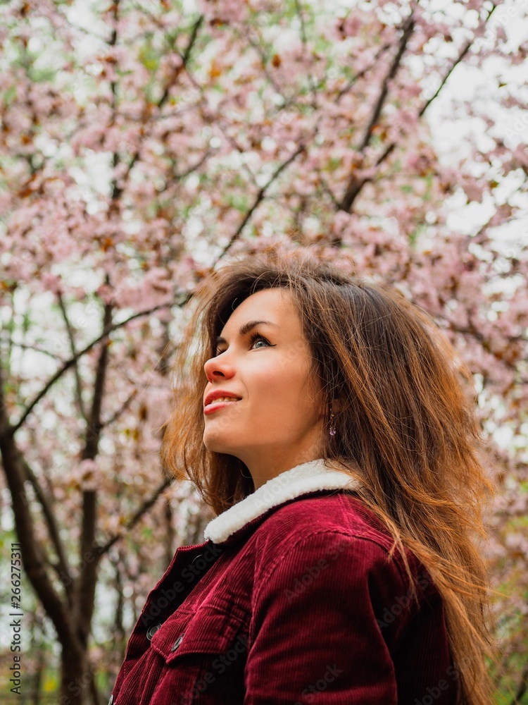 Brunette girl with long hair in a red jacket is surrounded by pink Sakura flowers Brunette girl with long hair in a red jacket is surrounded by pink Sakura flowers