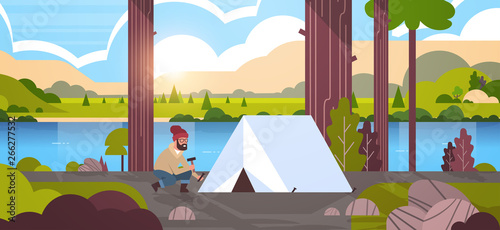 man hiker camper installing a tent preparing for camping hiking concept sunrise landscape nature river mountains background flat horizontal full length