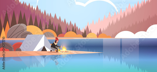woman hiker making fire near tent camp girl holding firewood for bonfire hiking camping concept autumn landscape nature river forest mountains background flat horizontal full length