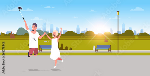 young couple using selfie stick taking photo on smartphone camera man woman jumping having fun city urban park sunrise landscape background full length horizontal