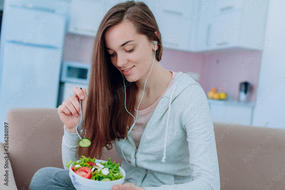 Young happy healthy woman in sportswear and in headphones eating fresh vegetable salad for snack after fitness exercises and workout at home. Balanced organic diet and clean eating. Young happy healthy woman in sportswear and in headphones eating fresh vegetable salad for snack after fitness exercises and workout at home. Balanced organic diet and clean eating.