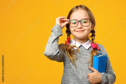 Portrait of a cute little kid girl on a yellow background. Child schoolgirl looking at the camera, holding a book and straightens glasses. The concept of education. Copy space. photo