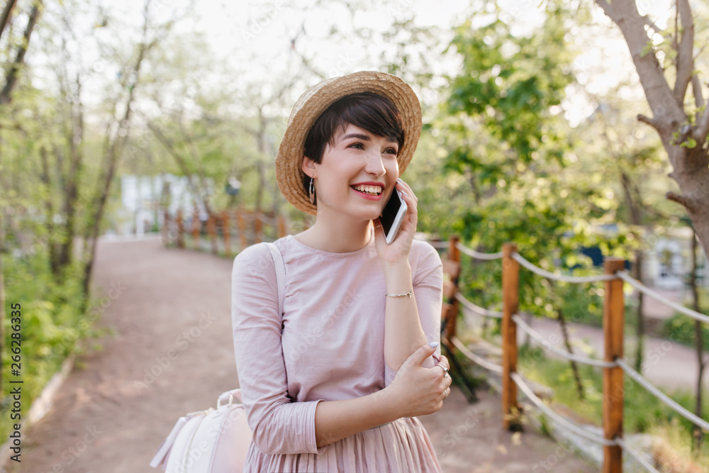 Amazing girl with short dark hair speaking on phone and looking up with smile. Outdoor portrait of inspired young lady wearing summer hat and cute gown walking down the alley.