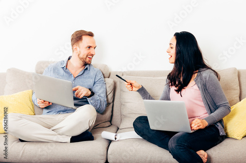 young happy couple sitting on a couch with laptop - freelancers, students, home connection, family lifestyle concepts