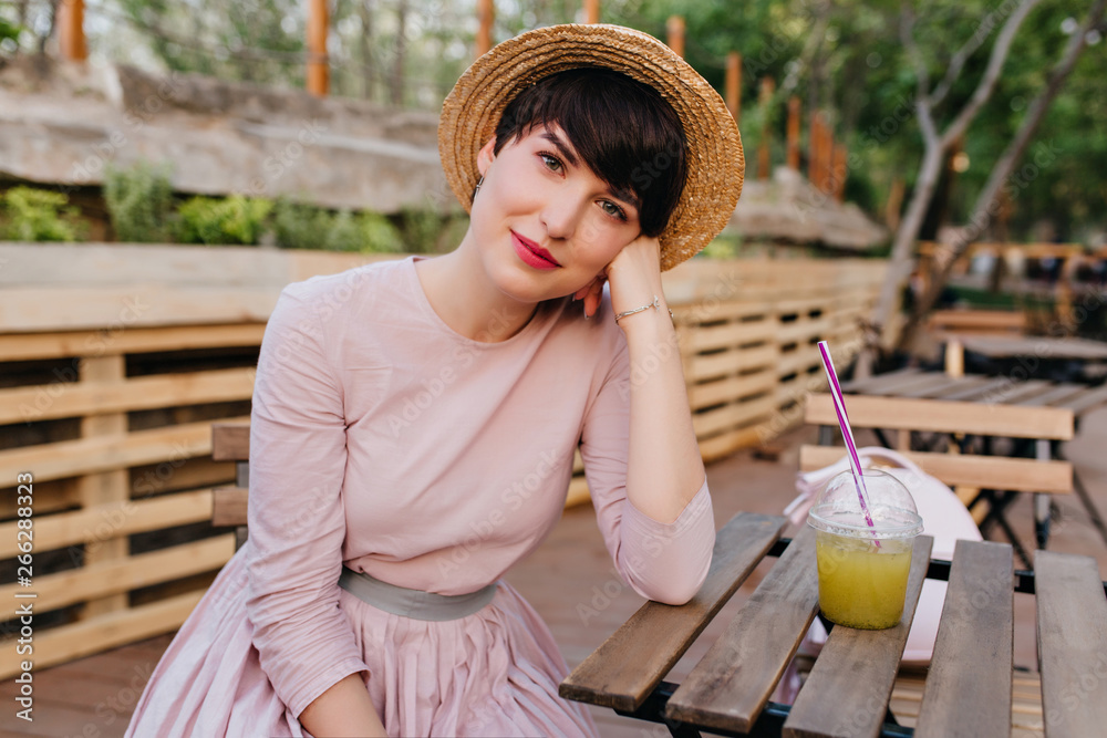 Obraz premium Lovely girl with nude makeup sitting at the wooden table propping face with hand. Portrait of charming young lady in straw hat and trendy pastel dress resting in outdoor cafe with glass of juice.