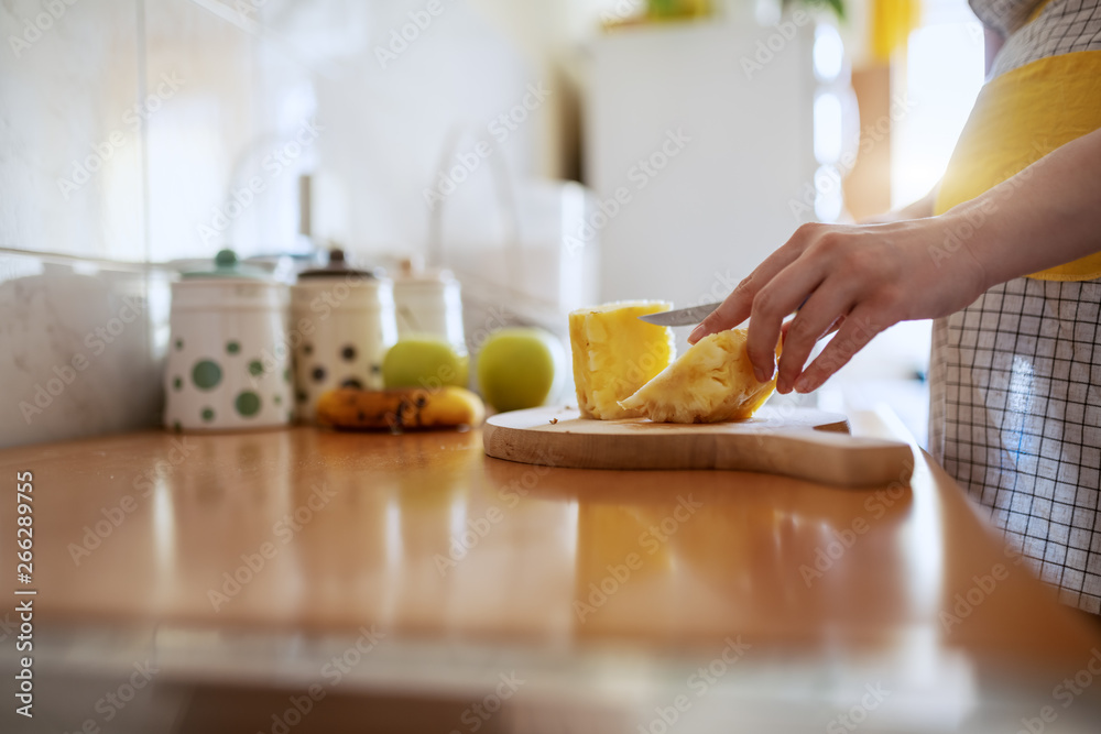 Caucasian pregnant woman wearing apron and cutting pineapple on cutting board in kitchen.