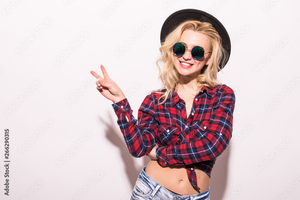 Smiling blonde woman in checkered shirt and hat showing peace gestures and looking at the camera over white background