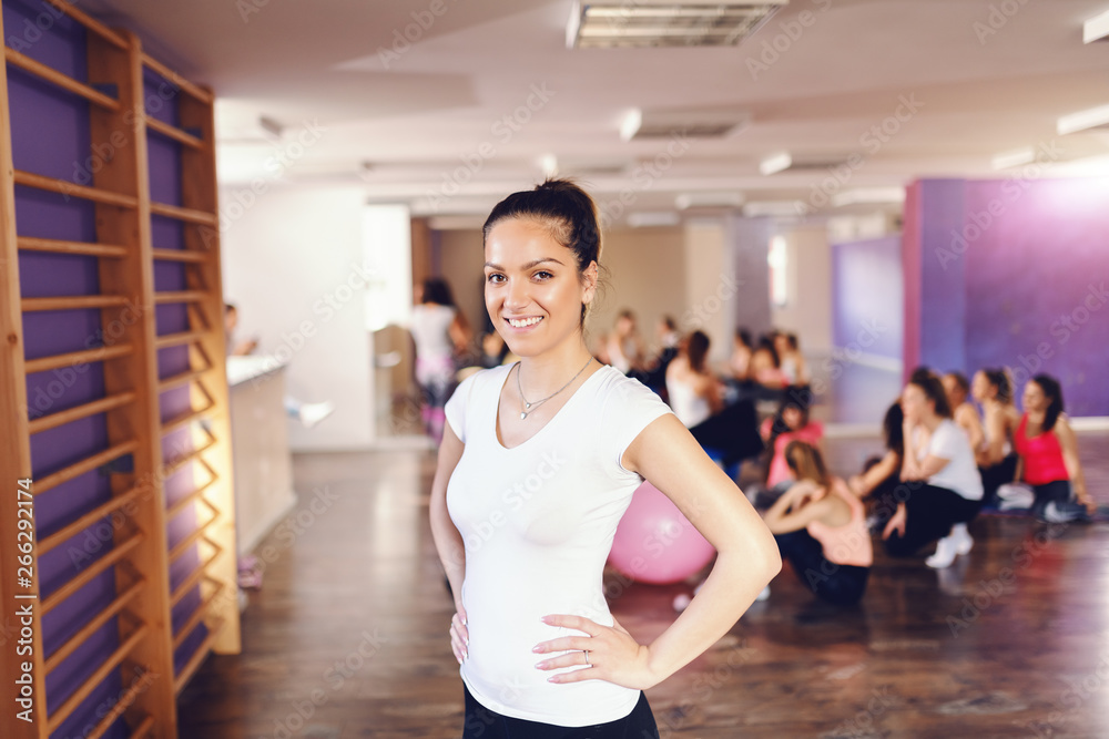 Young smiling Caucasian brunette in sportswear holding hands on hips, posing and looking at camera. In background women resting after exercises. Gym interior. Young smiling Caucasian brunette in sportswear holding hands on hips, posing and looking at camera. In background women resting after exercises. Gym interior.